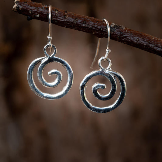 Et par Spiral Hængeøreringe 925s Sterling Sølv øreringe hængende fra en vandret brun gren. Øreringene har et åbent cirkulært design med et dominerende spiralmønster. Baggrunden er sløret, hvilket smukt understreger disse elegante finsk-inspirerede øreringe.