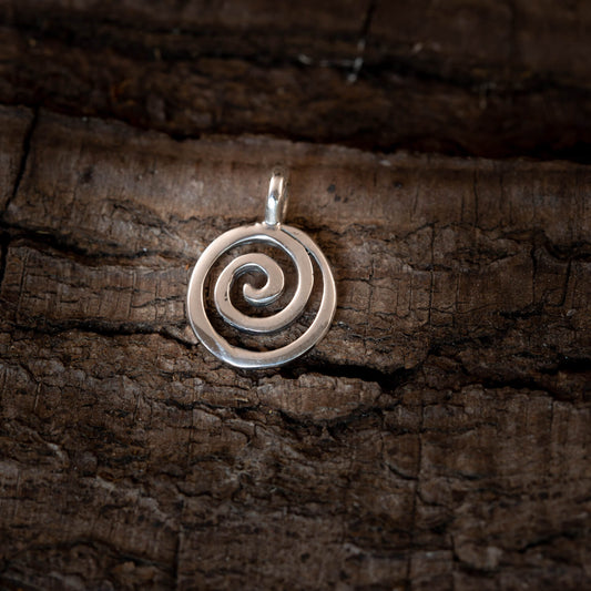 En Spiral Vedhæng 925s Sterling Sølv med et delikat spiralmotiv hviler på en tekstureret, mørk træoverflade. Vedhænget har et enkelt hvirveldesign, der sløjfer jævnt fra midten og udad. Dette stykke kunne sagtens være en udstilling på Finlands Nationalmuseum.