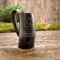 Indlæs billede i Gallery viewer, Et Vikingekrus Valknut Horn (600-800 ml) med nordiske symboler hviler på en træoverflade prydet med grønt mos og en plante i baggrunden, der fremkalder vikingearvens ånd.
