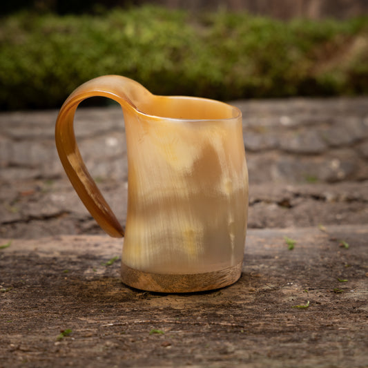 Ett handgjort mini-vikingahornkrus i naturfärg (100 ml) med ett slätt, böjt handtag står på en träyta. Kruset har ett naturligt, jordnära utseende med marmorerade beige och bruna toner. Bakgrunden är suddigt grön.