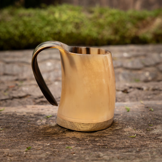 Ett Mini Vikingekrus Horn Nature (100 ml) med ett böjt handtag står på en träyta. Bakgrunden är suddig med gröna löv, vilket framhäver kannans rustika och eleganta design.