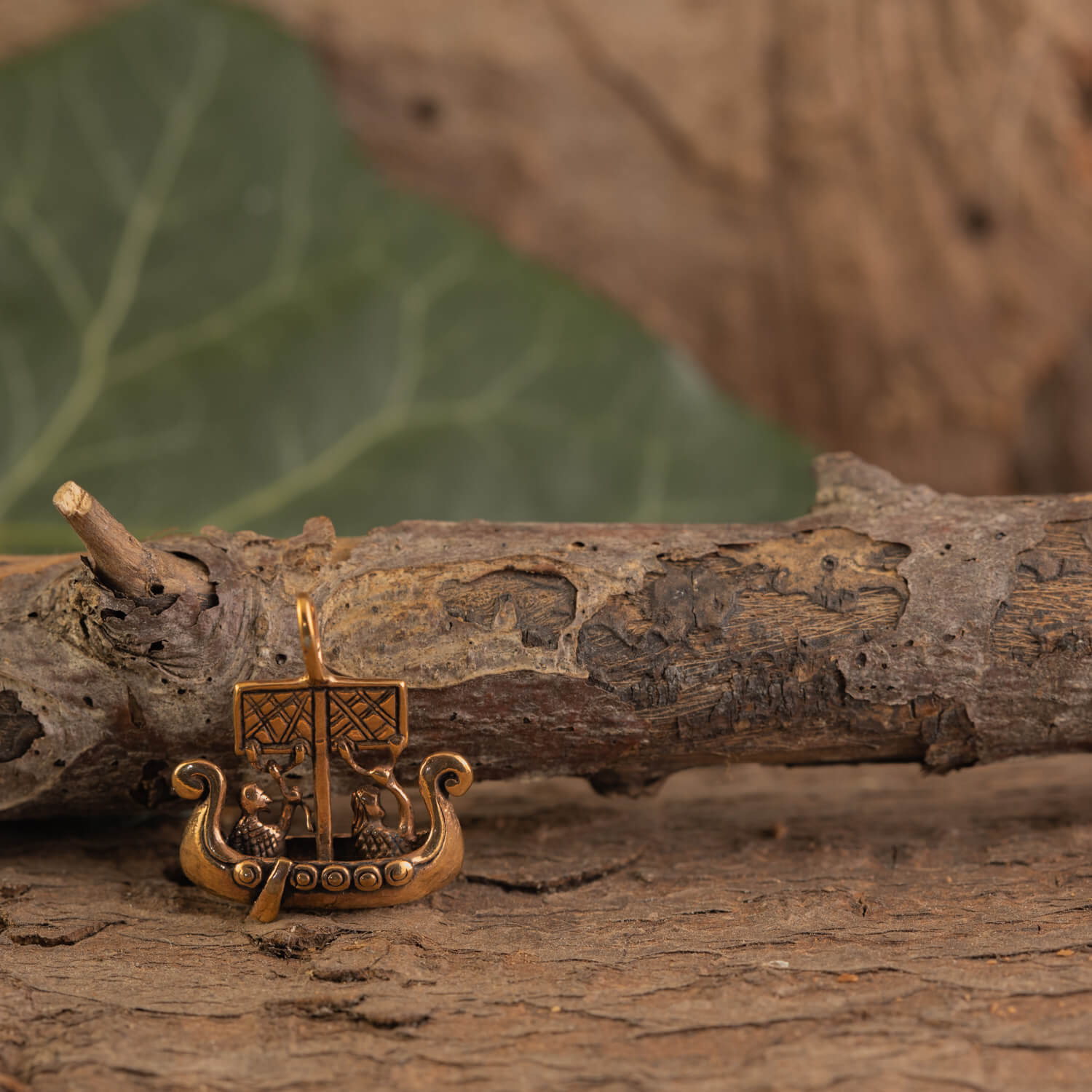 En lille figur, Vikingskib Vedhæng Bronze, hviler på en ru barkoverflade. Baggrunden har sløret grønt løv, der skaber en naturlig og rustik ramme for den indviklede miniaturebåd med detaljerede designs og buede ender.