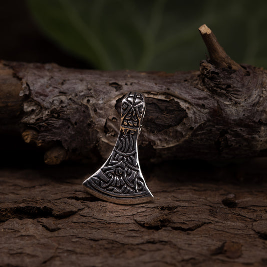 En liten, intrikat designad yxhuvudsberlock i 925s silver vilar på en strukturerad träyta, med suddig grön bakgrund. Vikingesmycket har detaljerade gravyrer och verkar vara inspirerat av nordiska eller vikingamotiv.