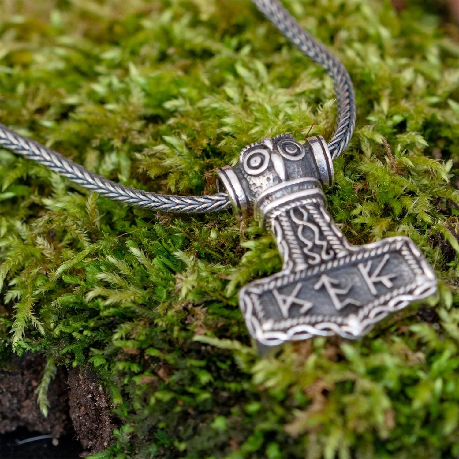 Et nærbillede af "Halskæde Rævehale 925s Sølv 3mm" vedhæng formet som Thors hammer (Mjolnir), prydet med indviklede designs og runer. Det middelalderinspirerede sterling sølv vedhæng drapererer elegant fra en flettet kæde, der hviler yndefuldt på en seng af levende grønt mos.