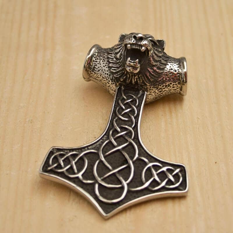 En smukt udformet Thors Hammer Vedhæng Stål, formet som Mjölnir, ligger på en træoverflade. Toppen af vedhænget viser en detaljeret udskæring af et brølende dyrehoved, mens selve hammeren er udsmykket med indviklet keltisk knudeværk, der fanger essensen af Vikingesmykker.