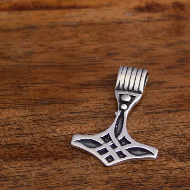 En Thors Hammer Vævet Mønster 925s Sølv formet som Thors Hammer (Mjölnir) med indviklede designs ligger på en træoverflade. Håndtaget har et rillet mønster, og hovedet har et unikt mønster i mørkere fordybninger.