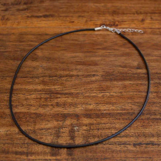 En enkel svart halskedja med ett litet silverspänne och förlängningskedja ligger platt på en träyta. Stålhalskedjan med svart coating är rund och kontrasterar vackert mot den strukturerade bruna bakgrunden.