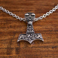 Indlæs billede i Gallery viewer, Et nærbillede af en sølvhalskæde med et vedhæng formet som Thors hammer (Mjölnir). Vedhænget har indviklede, indgraverede detaljer med nordisk inspireret design og er fastgjort til en robust Halskæde Ankerkæde i Stål 5mm. Den hviler elegant på en træoverflade.
