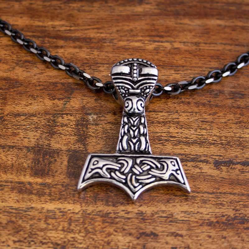 Et detaljeret vedhæng i form af Thors hammer, Mjölnir, hænger fra en Halskæde Ankerkæde Sort Stål 5mm. Vedhænget har indviklede, nordisk-inspirerede udskæringer og mønstre i en slående kontrastfarve. Den hviler på en træoverflade.