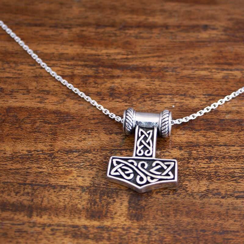 En sølv Halskæde Ankerkæde 925s Sølv 2 mm halskæde med et vedhæng, der er indviklet designet som Thors hammer (Mjölnir) med keltiske knudemønstre. Ægte 925s Sterling sølv vedhæng hænger i en løkke med lignende design og hviler på en træoverflade. Fås i forskellige længder på en ankerkaede.